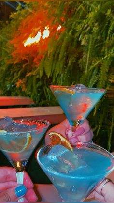 two people holding up martini glasses with blue liquid and orange slices on the rims