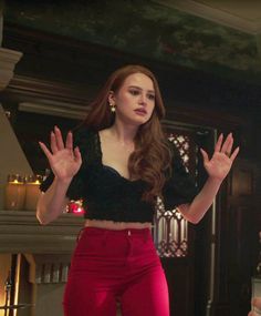 a woman standing in front of a fireplace wearing red pants and black cropped top