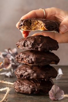 a hand holding a chocolate covered doughnut in front of a stack of other donuts