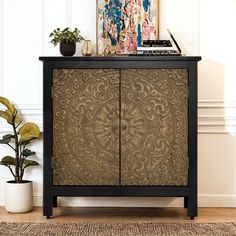 a black cabinet with an intricate design on the front and sides, next to a potted plant