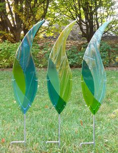 three green and blue glass leaves on metal stands in the grass with trees in the background