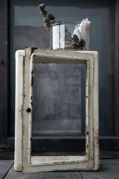 an old white cabinet with a glass vase on it's top sitting on a wooden floor