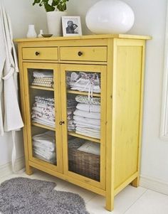 a yellow cabinet with baskets and towels on it next to a white vase filled with flowers