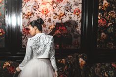 a woman standing in front of a wall with flowers on it and wearing a white dress