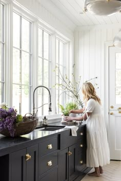 a woman standing in front of a kitchen sink next to a counter with flowers on it