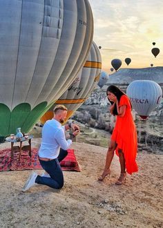 a man kneeling down next to a woman in an orange dress near hot air balloons