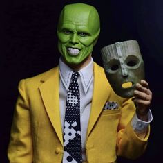 a man in a yellow suit and tie holding a mask with his right hand up to the camera