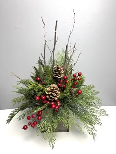 a christmas arrangement with pine cones, berries and greenery in a square vase on a table