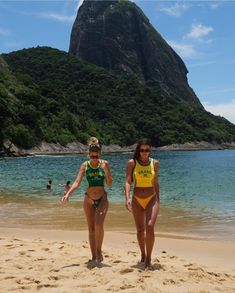 two women in bikinis standing on the beach next to an island with a mountain behind them