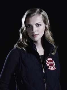 a woman with long blonde hair wearing a black jacket and red badge on her chest