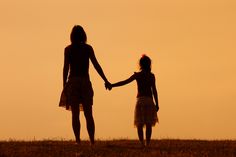 two women holding hands while standing on top of a grass covered hill in the sunset