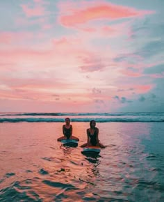 two people are sitting on surfboards in the water at sunset or dawn, with pink clouds above them