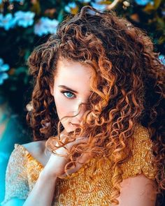 a woman with curly hair and blue eyes is posing for a photo in front of some flowers