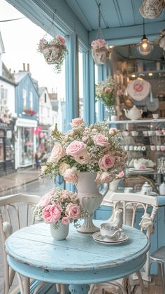 a blue table with pink flowers on it in front of a storefront and some chairs