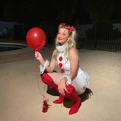 a woman dressed up as a clown holding a red balloon in her hand and sitting on the ground