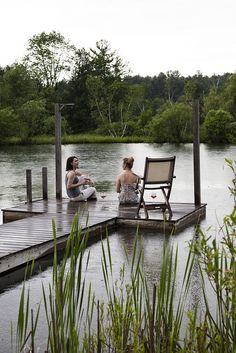 two women sitting on a dock next to a body of water with trees in the background