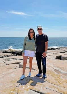 a man and woman standing on rocks near the ocean with their arms around each other