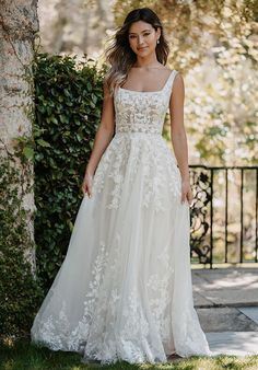 a woman wearing a wedding dress standing in front of a tree and bushes with her hand on her hip