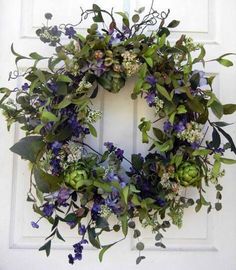 a wreath with purple flowers and green leaves hanging on a white front door, surrounded by greenery