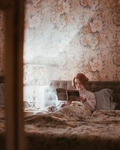 a woman laying in bed reading a book while looking at the camera through a window