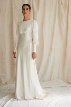 a woman standing in front of a white backdrop wearing a long sleeved wedding dress