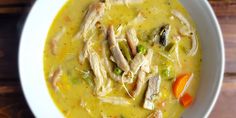 a white bowl filled with chicken and vegetable soup on top of a wooden table next to a spoon