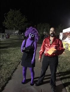 two people in costumes standing on a sidewalk at night with one person wearing purple hair and the other holding a yellow envelope