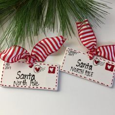 two red and white striped christmas ornaments hanging from a pine tree with santa north pole tags on them