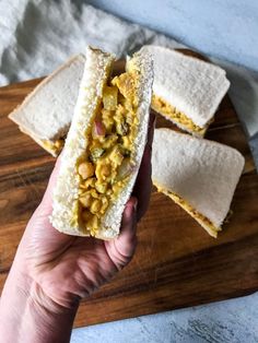 a person holding up a sandwich on top of a wooden cutting board next to two pieces of bread