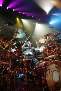 a man playing drums in front of colorful lights on stage with his hands up to the drum sticks