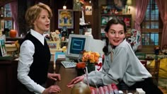 two women sitting at a desk in front of a computer monitor and keyboard, talking to each other