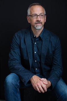 a man with glasses and a beard is sitting on a black chair in front of a dark background