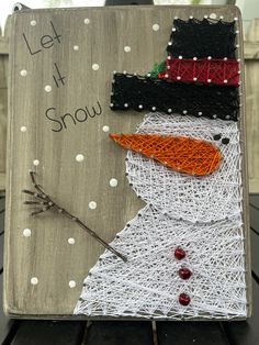 a snowman made out of yarn sitting on top of a wooden sign that says let it snow