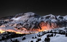 a snowy mountain with trees and lights in the distance at night, surrounded by snow covered mountains