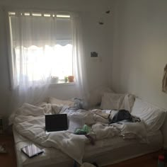 an unmade bed sitting in front of a window next to a laptop on top of a wooden floor