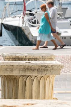 two people walking past a boat docked in the water near a stone pillar with an ornate design on it