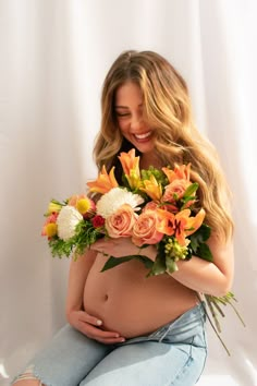 a pregnant woman with flowers on her belly sitting in front of a white backdrop holding a bouquet