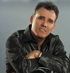 a man with his arms crossed leaning on a motorcycle and looking at the camera while wearing a black leather jacket