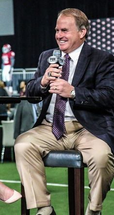 a man in a suit and tie sitting on a chair holding a microphone while talking to someone