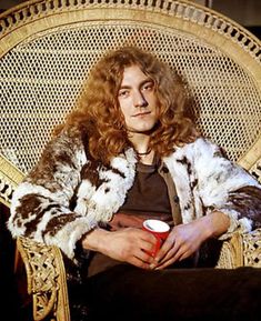 a man with long hair sitting in a chair and holding a coffee cup while wearing a fur coat