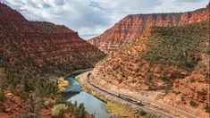 a train traveling through a canyon next to a river in the middle of it's mountains