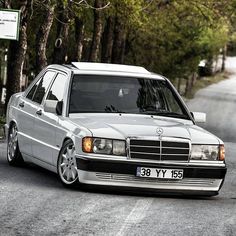 a white mercedes benz is parked on the side of the road in front of some trees