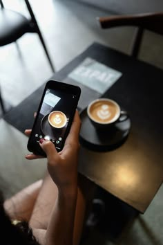 a person holding up a smart phone to take a photo with two cups of coffee on the table