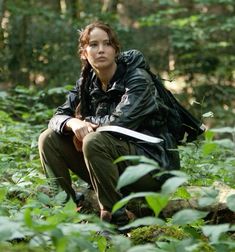 a woman crouching down in the woods with a book on her lap and looking at the camera