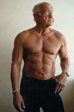 an older man with no shirt standing in front of a wall and posing for the camera