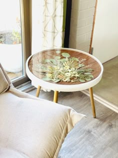 a glass table with flowers on it sitting in front of a window next to a couch