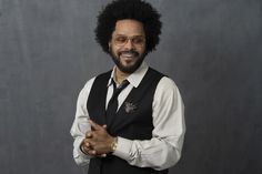 a man with an afro wearing glasses and a black vest is standing in front of a gray background