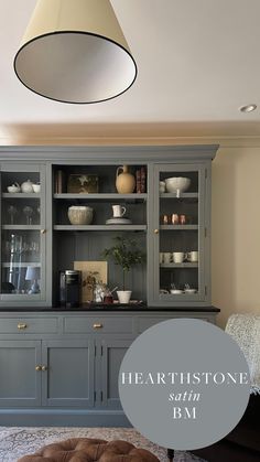 a gray china cabinet with glass doors and shelves in the middle, next to a brown ottoman