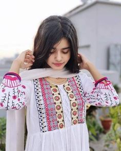 a woman wearing a white dress with colorful designs on her shoulders and arms, standing in front of a house