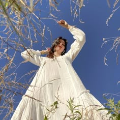 a woman in a white dress is standing among tall grass and trees with her arms outstretched